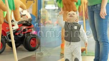 两岁可爱的金发男孩在商店门口的商场里向妈妈展示大软玩具